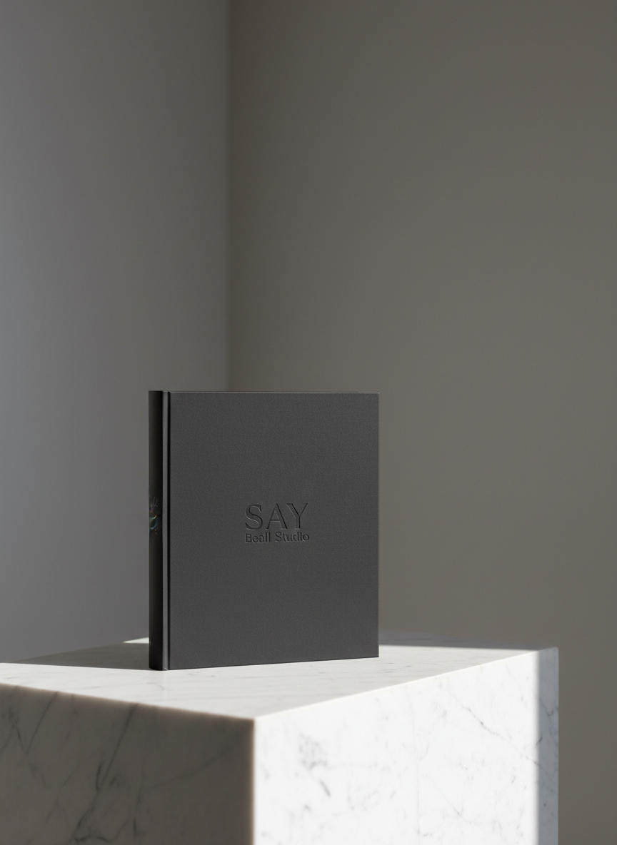 A single, impeccably crafted hardcover portfolio book in deep matte charcoal, embossed with a subtle, tone-on-tone SAY Beall Studio logotype at the center. The book rests on a low, wide plinth of honed white marble, set against a soft, blurred background of neutral gray walls and a faint architectural corner line. Cool, overcast daylight falls from the right, creating a delicate rim of light along the book’s spine and casting a refined shadow onto the marble. Shot at eye level with a slightly off-center, rule-of-thirds composition, the image emphasizes negative space and refined materiality. The photographic realism and clean, modern aesthetic communicate sophistication, permanence, and design as enduring communication rather than fleeting trend.
