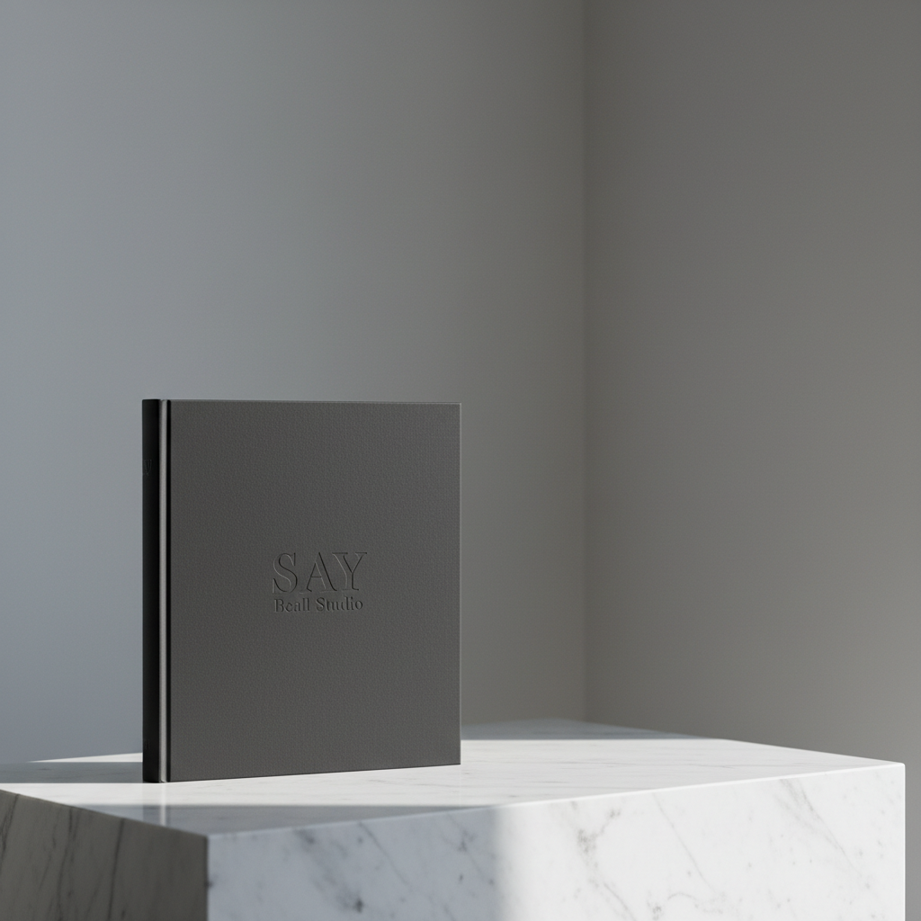 A single, impeccably crafted hardcover portfolio book in deep matte charcoal, embossed with a subtle, tone-on-tone SAY Beall Studio logotype at the center. The book rests on a low, wide plinth of honed white marble, set against a soft, blurred background of neutral gray walls and a faint architectural corner line. Cool, overcast daylight falls from the right, creating a delicate rim of light along the book’s spine and casting a refined shadow onto the marble. Shot at eye level with a slightly off-center, rule-of-thirds composition, the image emphasizes negative space and refined materiality. The photographic realism and clean, modern aesthetic communicate sophistication, permanence, and design as enduring communication rather than fleeting trend.